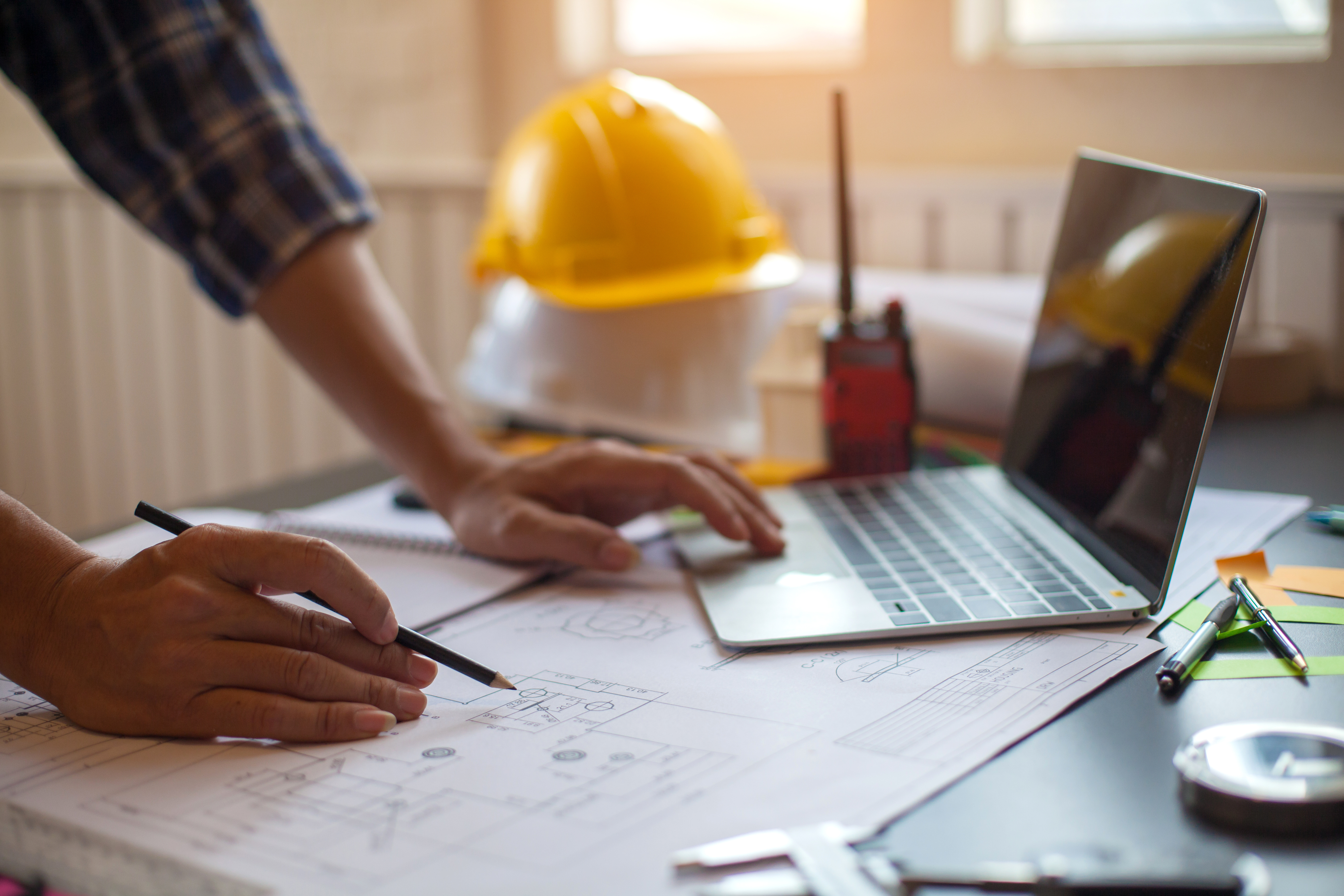 Construction worker reviewing blueprints at desk with laptop and hard hat